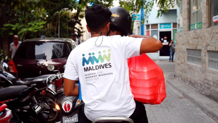 two people on a motorbike with a bag of meal packs. One of them is wearing a t-shirt with the Mission for Migrant Workers logo